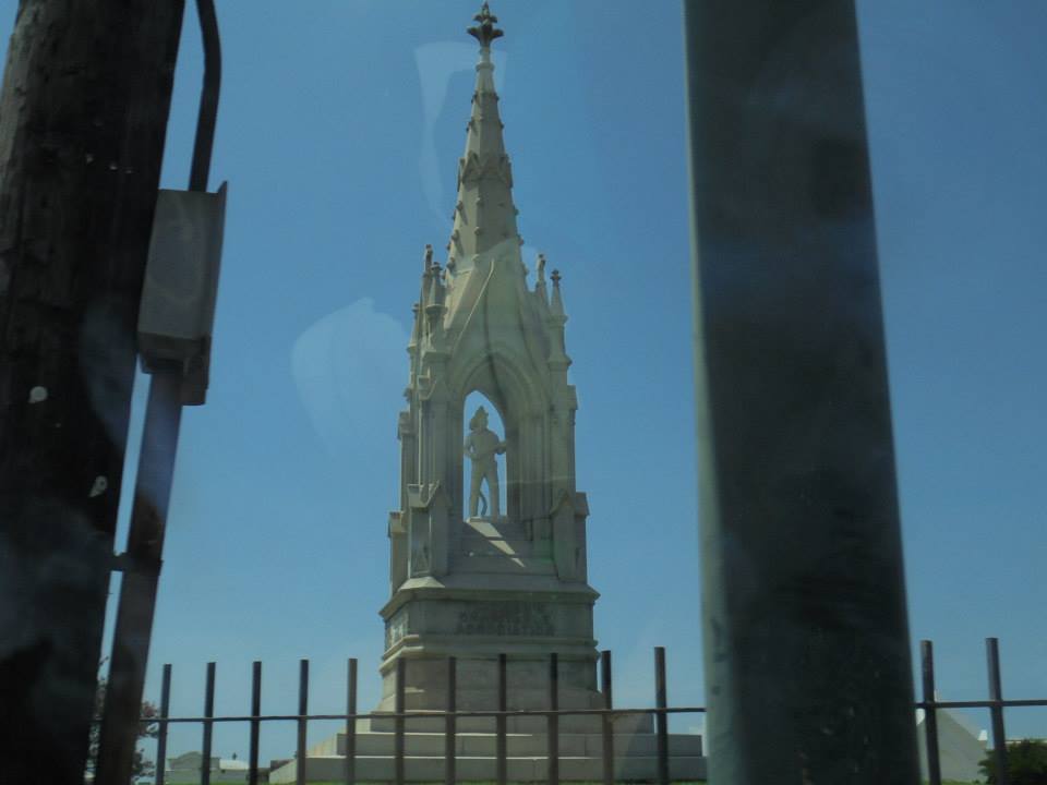 A tall, neo-Gothic stone monument rising from a grassy mound in Greenwood Cemetery, topped with a marble statue of a volunteer fireman. The tower is carved with pointed arches and decorative details, standing prominently against the sky. This memorial honors 19th-century New Orleans volunteer firefighters.