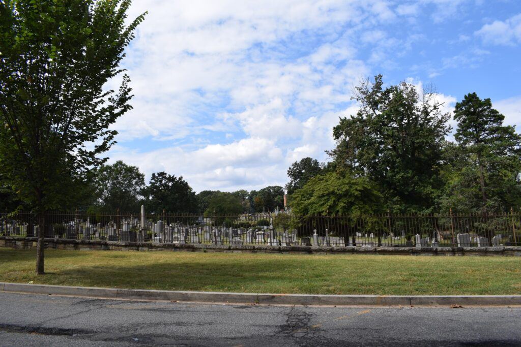 View of the United States Soldiers’ and Airmen’s Home National Cemetery from the roadside, showing rows of Civil War-era headstones stretching across a hill beneath large shade trees.