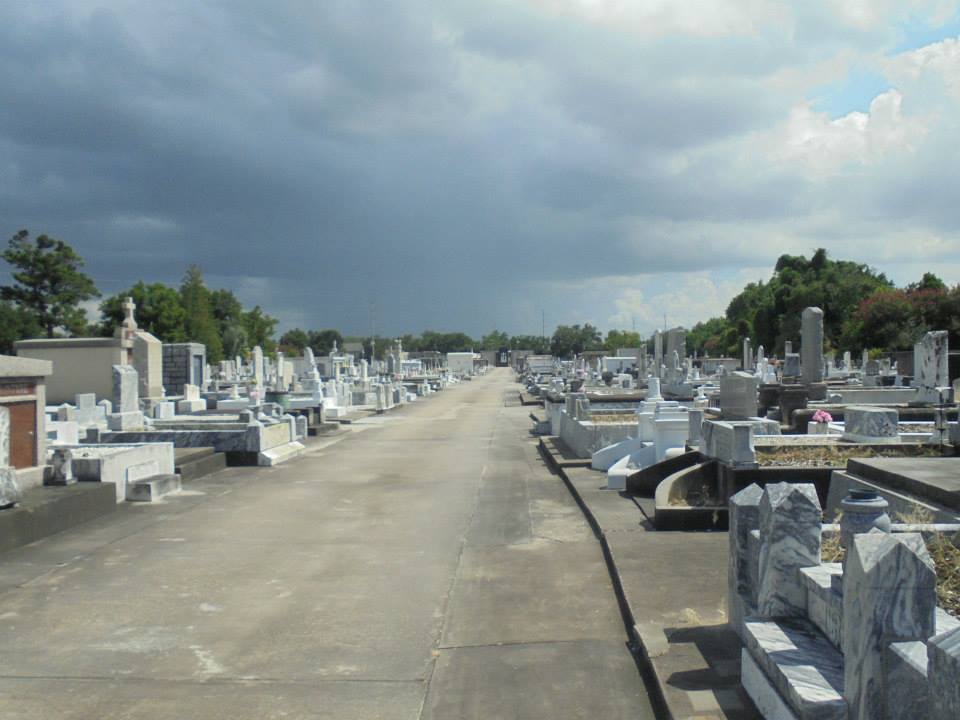 A wide view down the central pathway of St. Patrick Cemetery No. 2 in New Orleans, lined with rows of white marble tombs and coping graves under a bright summer sky.