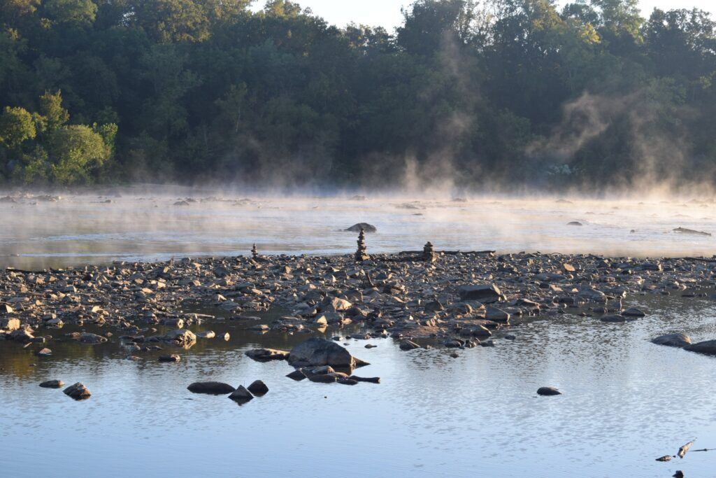 Fog drifting over the Potomac River at sunrise, soft golden light illuminating the still water and rocky shoreline.