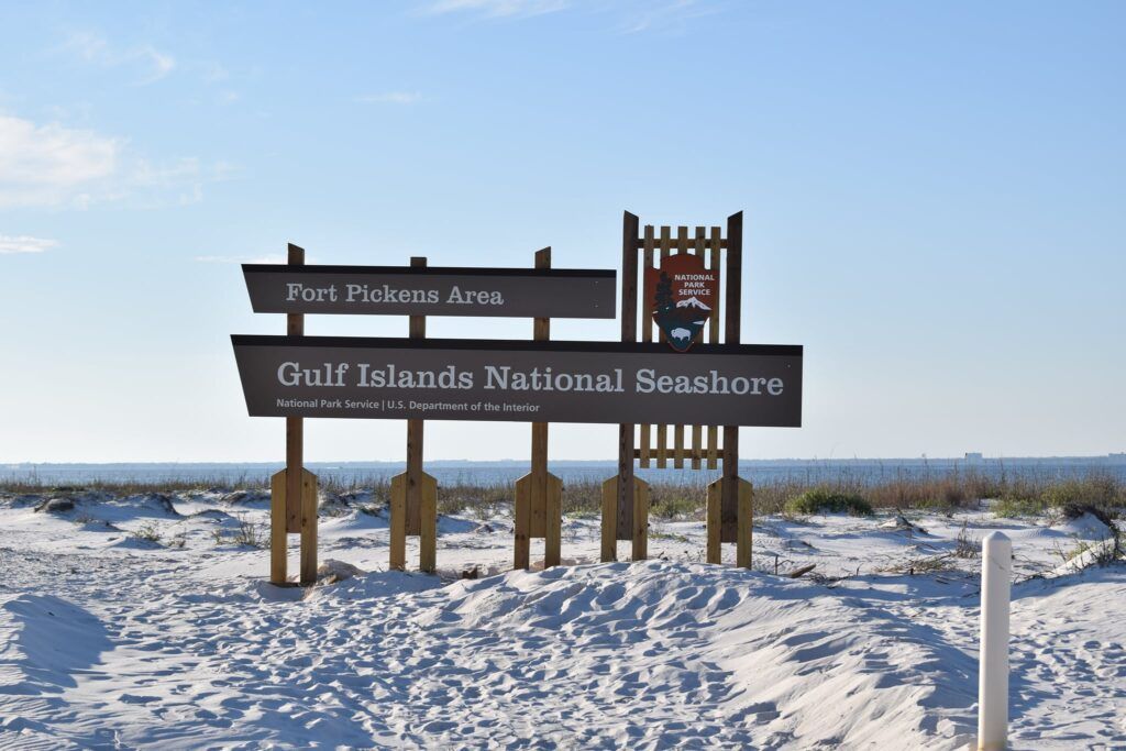 Entrance sign for Gulf Islands National Seashore Fort Pickens Area on top of white sand dunes with the crystal blue Gulf of Mexico in the background.