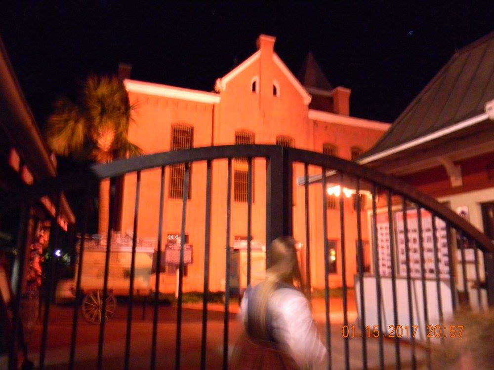Front exterior of the Old Jail in St. Augustine, Florida — pastel pink Romanesque Revival structure with barred windows and palm trees.