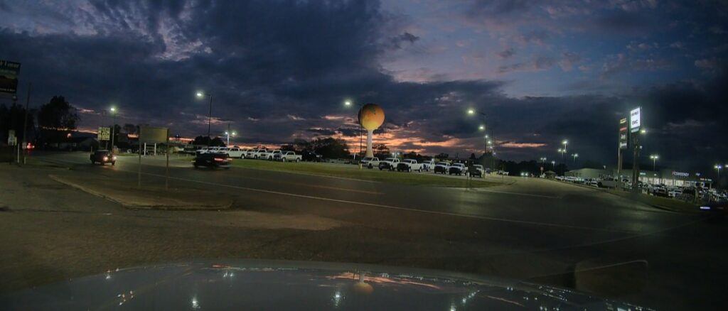 A glowing peach-shaped water tower rises over a gas station at dusk in Clanton, Alabama, its bright orange orb set against a purple-and-gold sky.