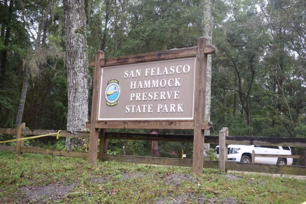 Entrance sign for San Felasco Hammock Preserve State Park in Gainesville, Florida, standing in front of dense hardwood forest with caution tape visible near the trail area.