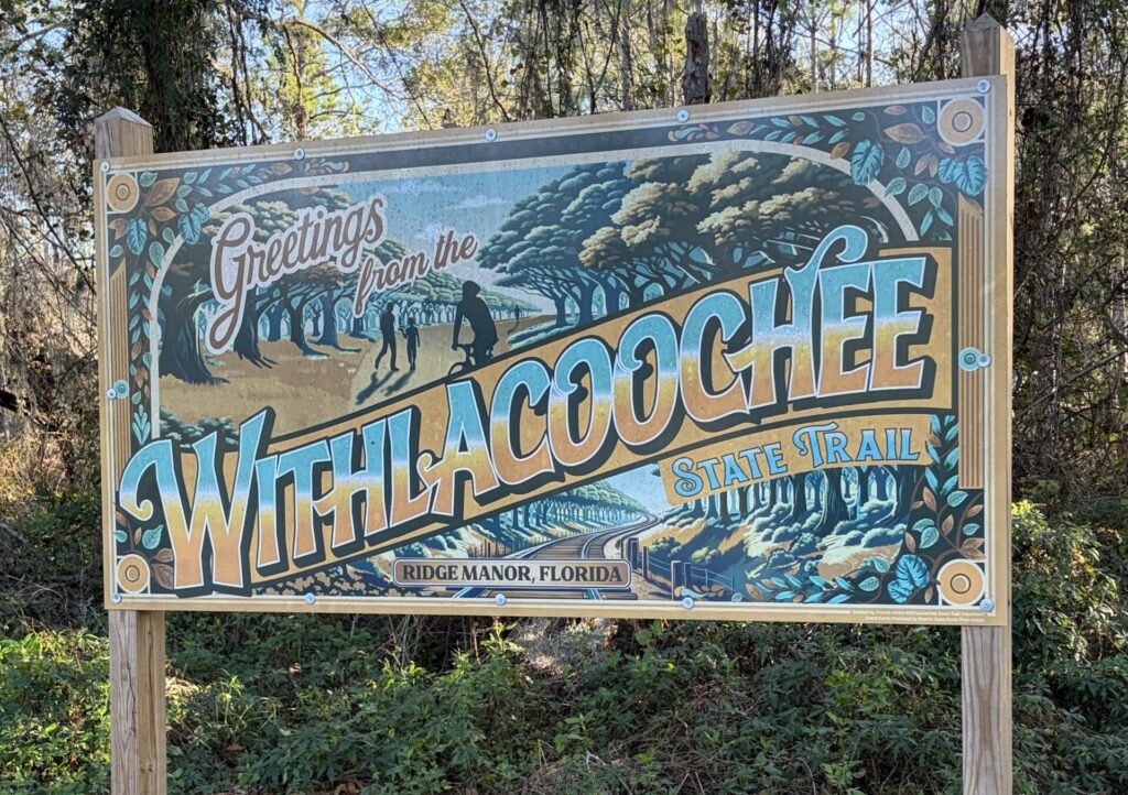 Illustrated sign reading “Greetings from the Withlacoochee State Trail” at Ridge Manor, Florida, marking the modern trail that follows a former railroad corridor.