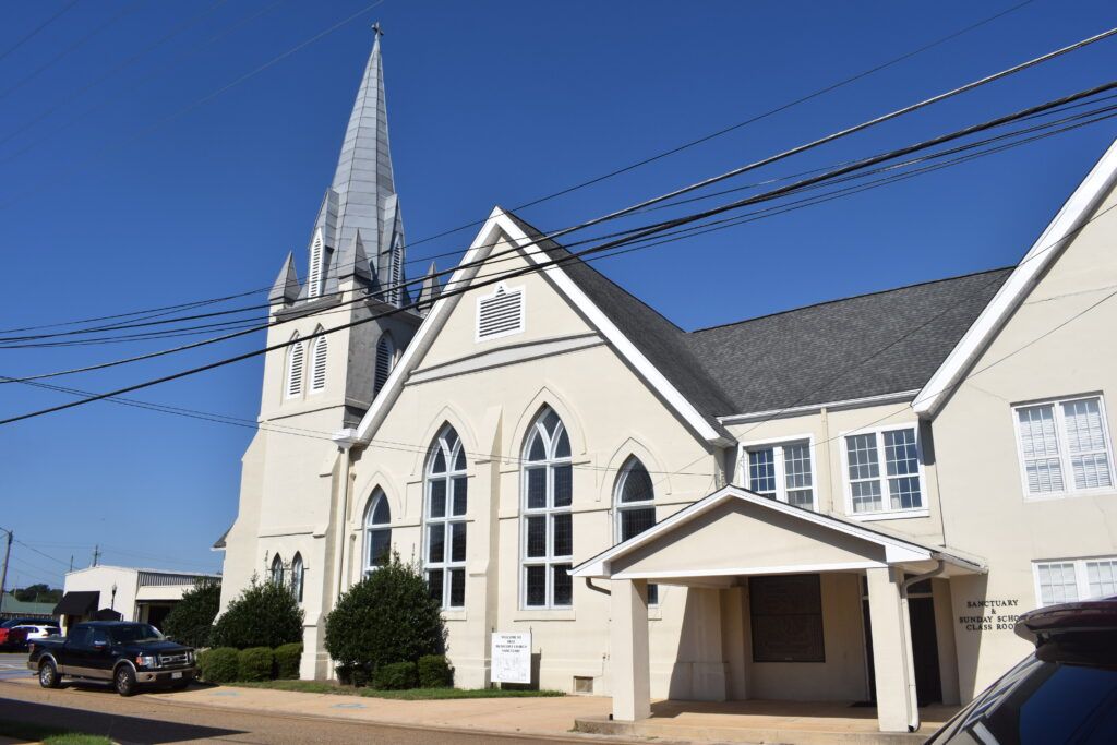 Exterior view of First Methodist Church in Enterprise, Alabama, showing the historic Gothic-style church with tall steeple and arched stained-glass windows under a clear blue sky.