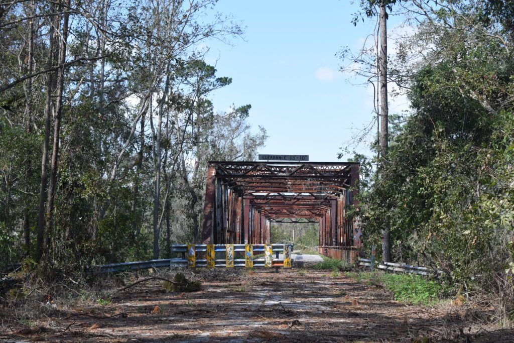 View down the length of the Hillman Bridge showing cracked asphalt, metal railings, and steel truss structure leading toward dense forest.