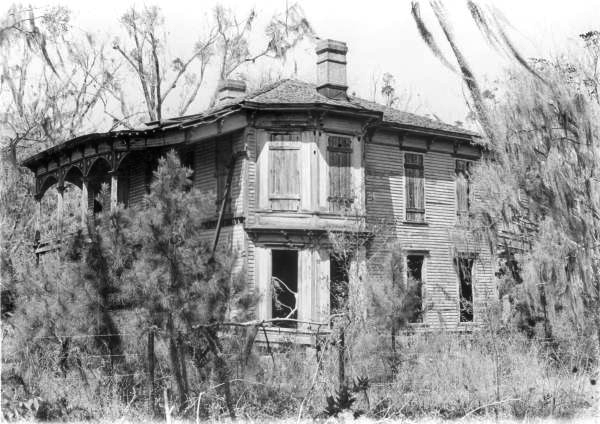 Black-and-white image of the Drew Mansion in Ellaville after abandonment, with damaged shutters, open doorways, and surrounding vegetation reclaiming the structure.