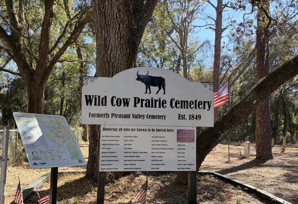 Entrance sign for Wild Cow Prairie Cemetery in Sumter County, Florida, displaying the cemetery name, establishment date of 1849, and a list honoring known burials, with oak trees and historic graves visible behind it.