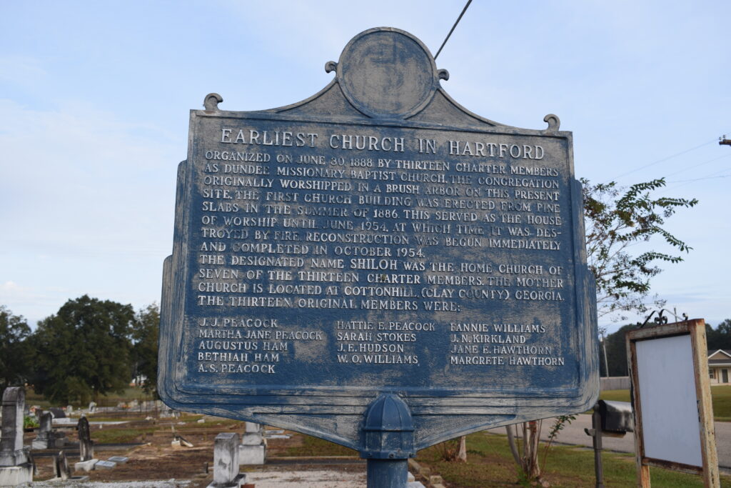 Weathered blue historical marker at Shiloh Baptist Church in Hartford, Alabama, displaying the names of the thirteen original members, standing before the quiet cemetery they helped shape.