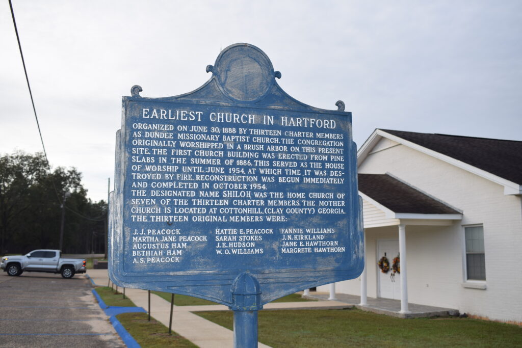 Weathered blue historical marker at Shiloh Baptist Church in Hartford, Alabama, telling the story of the town’s earliest church as it stands quietly beside the modern white chapel.