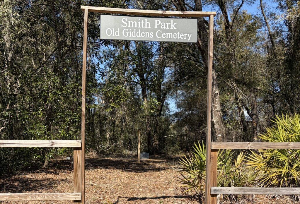 Entrance sign for Old Giddens Cemetery at Smith Park in Webster, Florida, with wooded area and no visible graves beyond the gate