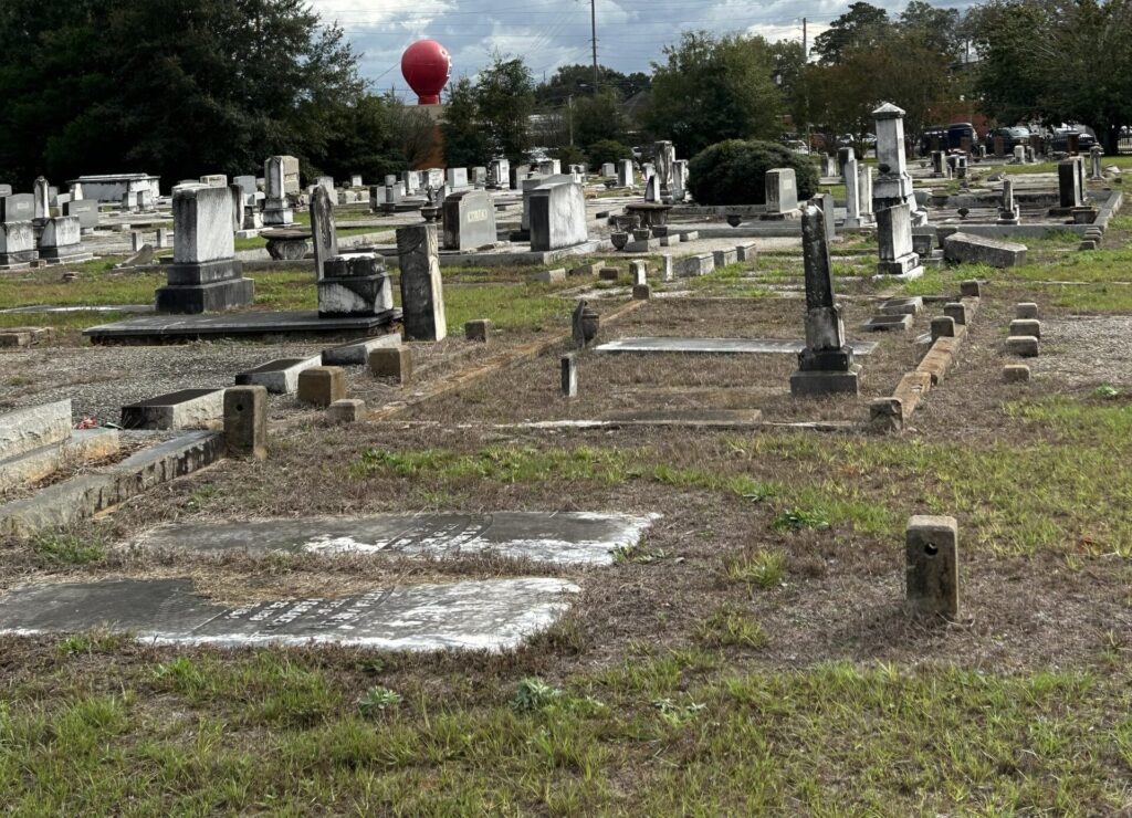 Wide view of Enterprise City Cemetery in Alabama with rows of weathered gravestones and monuments under cloudy skies, with a red water tower visible in the distance.