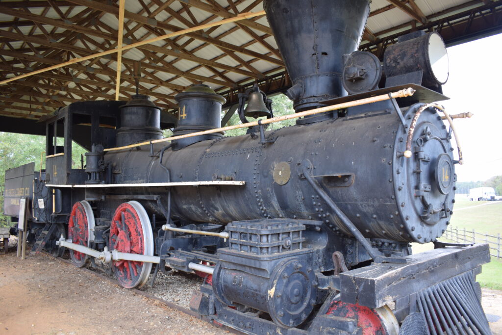 Historic 1881 steam locomotive on display at Pioneer Museum of Alabama in Troy, featuring a black engine body, red wheels, and detailed ironwork under an open-sided shelter.