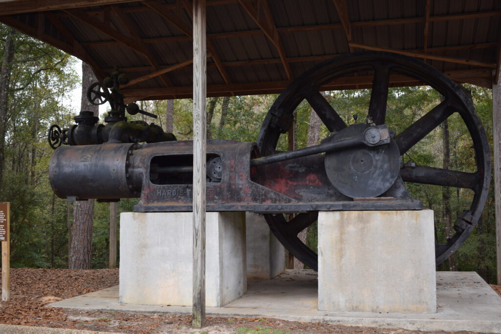 Historic steam engine on display at Pioneer Museum of Alabama in Troy, Alabama, featuring a large flywheel and iron machinery mounted on concrete supports beneath a wooden shelter surrounded by trees.