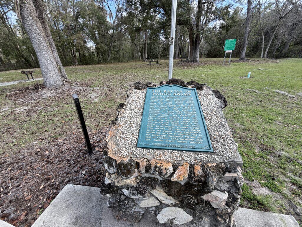 Close-up of the Battle of Wahoo Swamp historical marker on a stone base in a wooded Florida park, with trees, picnic tables, and grassy clearing in the background