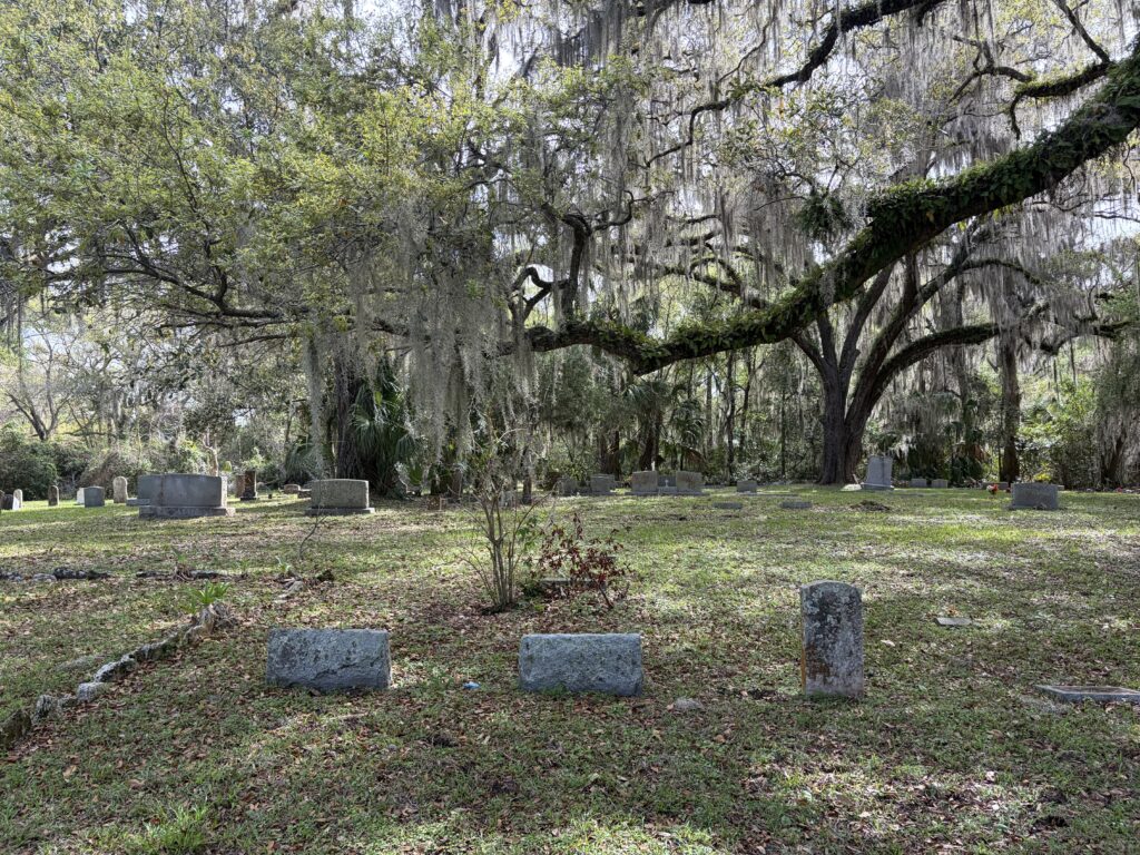 Scattered and weathered gravestones in Dampier Cemetery, Inverness, Florida beneath moss-draped live oak branches, reflecting the resting place of forgotten and unnamed individuals.