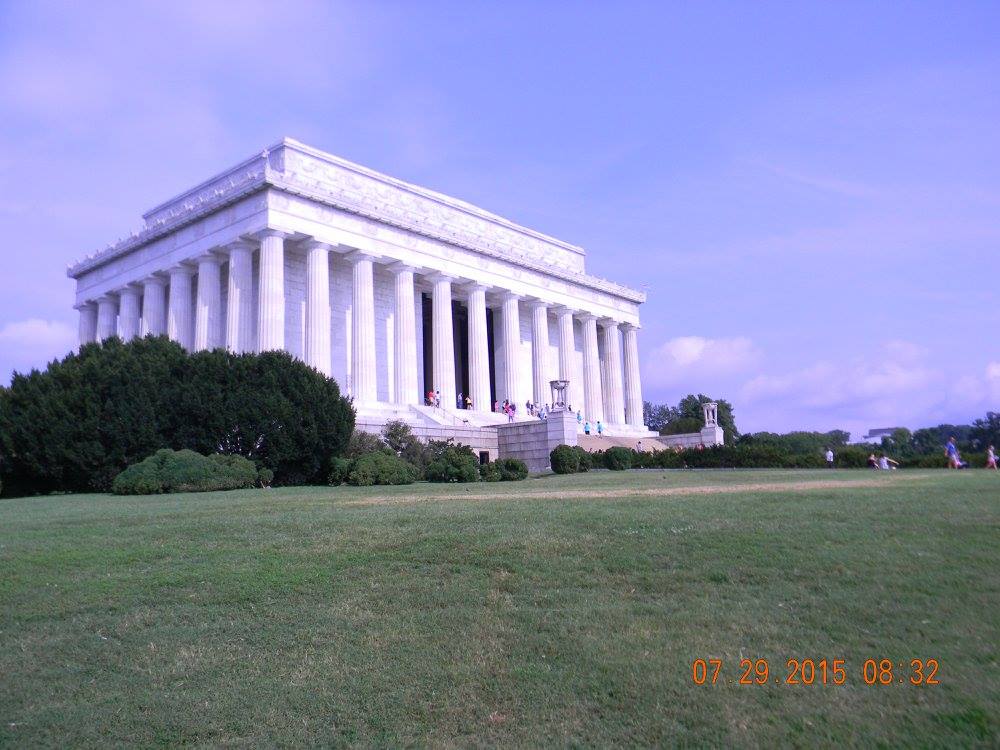 Wide view of the Lincoln Memorial in Washington, D.C., seen from the grassy lawn below under a hazy summer sky.