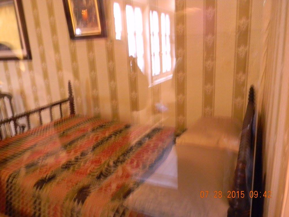 Historic bedroom inside the Petersen House showing the narrow bed where Abraham Lincoln was laid, with patterned wallpaper, a window letting in soft light, and simple 19th-century furnishings.
