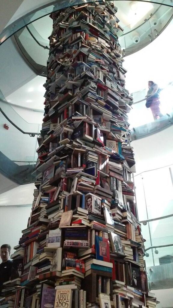 Towering column of stacked books about Abraham Lincoln rising through multiple floors inside the Petersen House museum, with visitors visible on an upper-level walkway.
