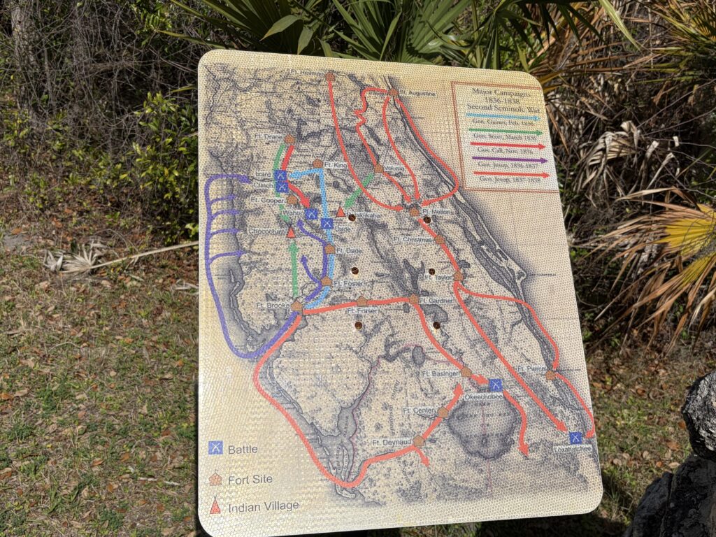 Interpretive map display showing military campaigns, forts, battles, and Seminole villages during the Second Seminole War in Florida, positioned beside dense palmettos and brush near Fort Armstrong.
