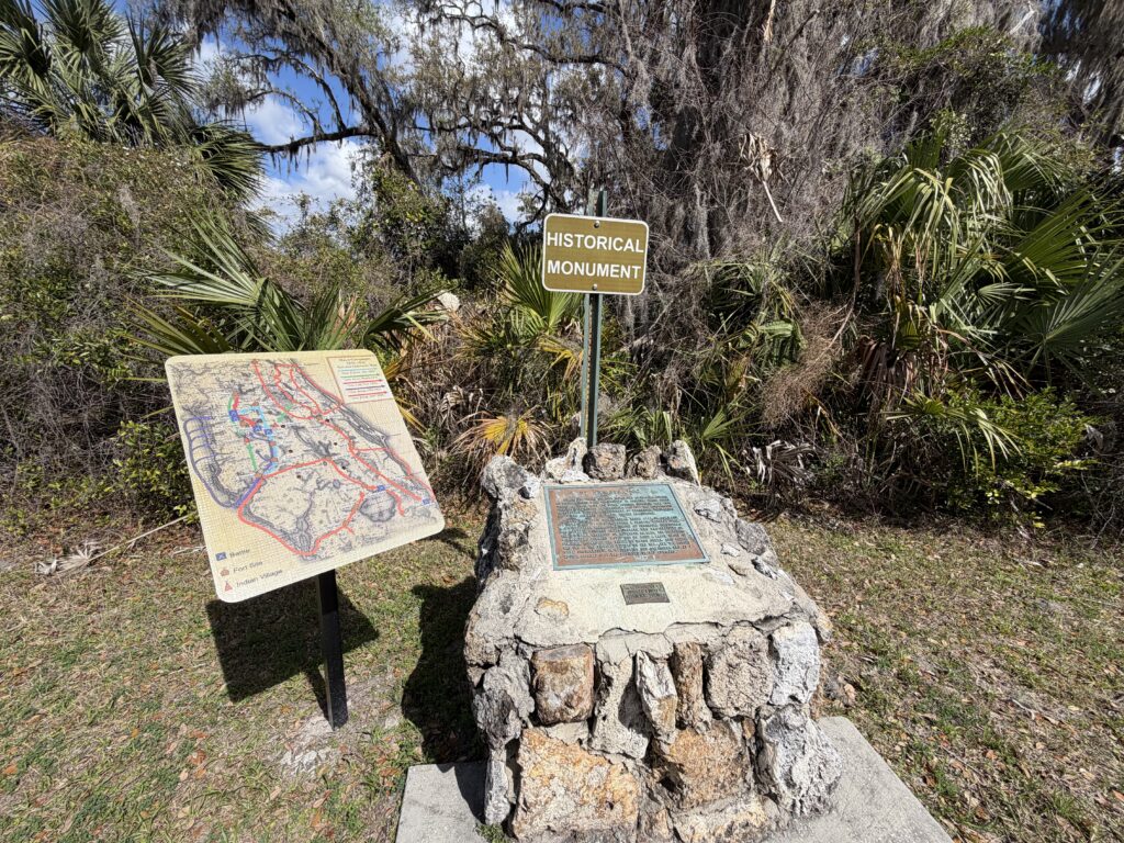 Historic monument and interpretive map marking the site of Fort Armstrong near Dade Battlefield in Florida, surrounded by palmettos, moss-draped oaks, and dense Florida wilderness beneath a bright blue sky.