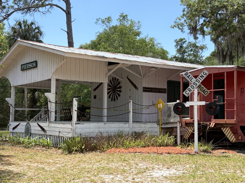 Historic Pierson Railroad Depot and red railcar display at Barberville Pioneer Settlement in Florida with a railroad crossing sign in the foreground.