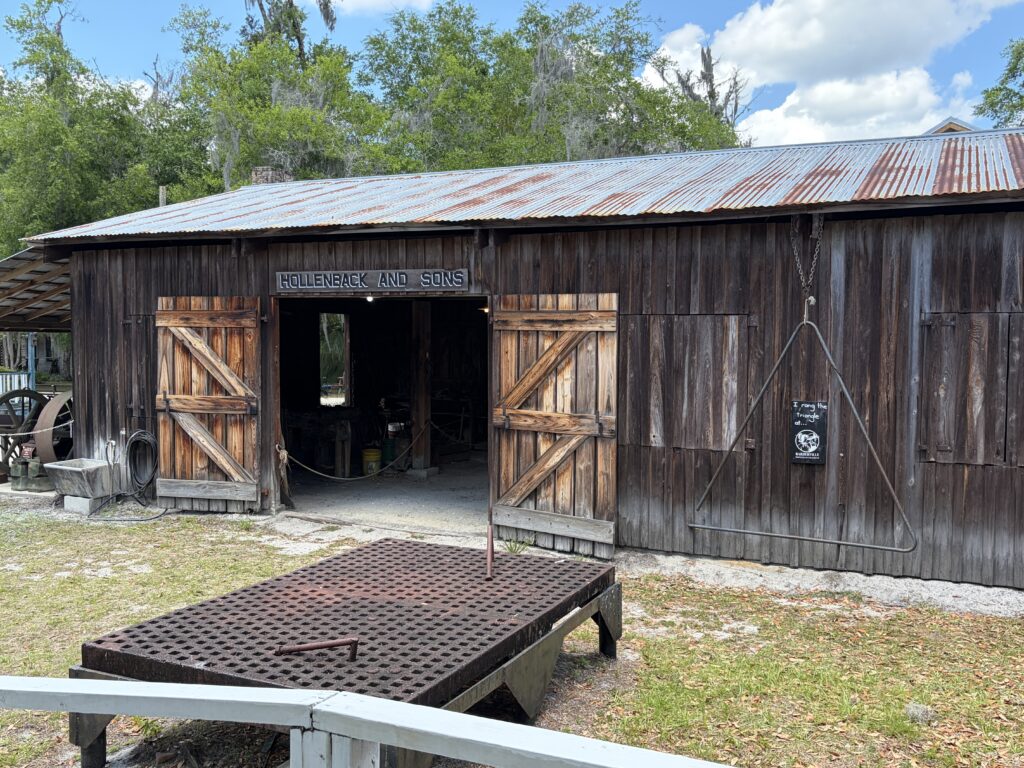 Historic blacksmith shop with weathered wooden walls and open barn doors at Barberville Pioneer Settlement in Florida, with an iron triangle hanging in the foreground.