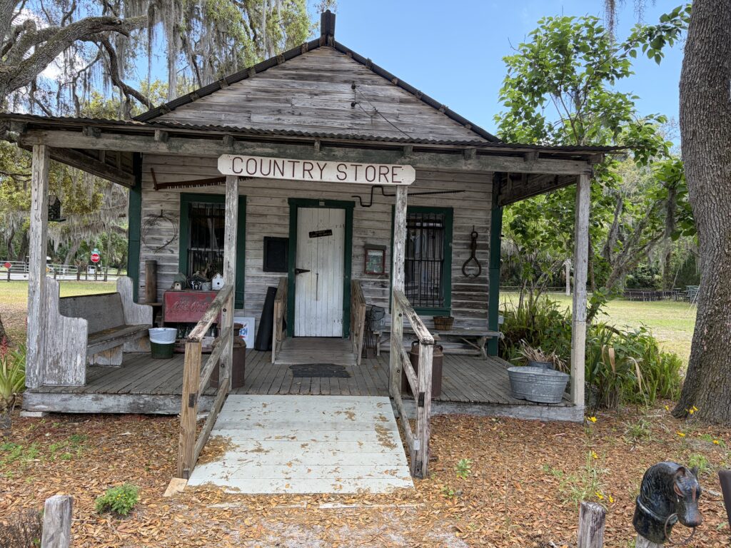 Weathered historic country store building with wooden porch and faded signage at Barberville Pioneer Settlement in Florida beneath moss-covered oak trees.