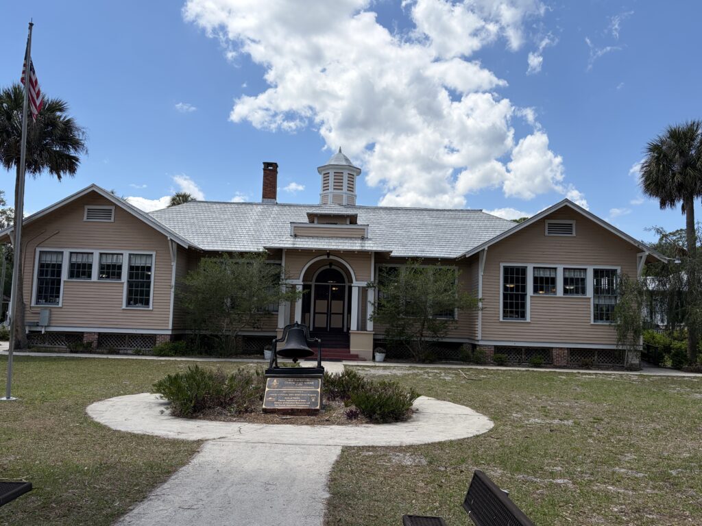 Historic Barberville Central High School building at Barberville Pioneer Settlement in Florida beneath a bright blue sky with large white clouds and an old school bell in the foreground.