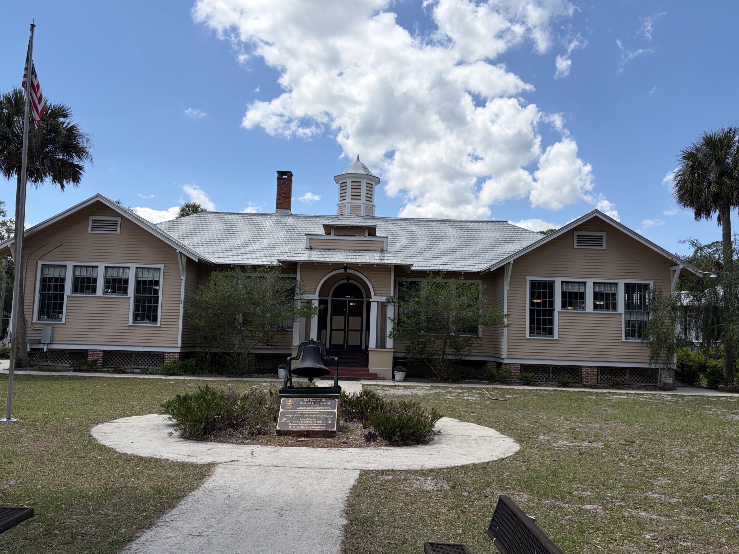 Historic Barberville Central High School building at Barberville Pioneer Settlement in Florida beneath a bright blue sky with large white clouds and an old school bell in the foreground.