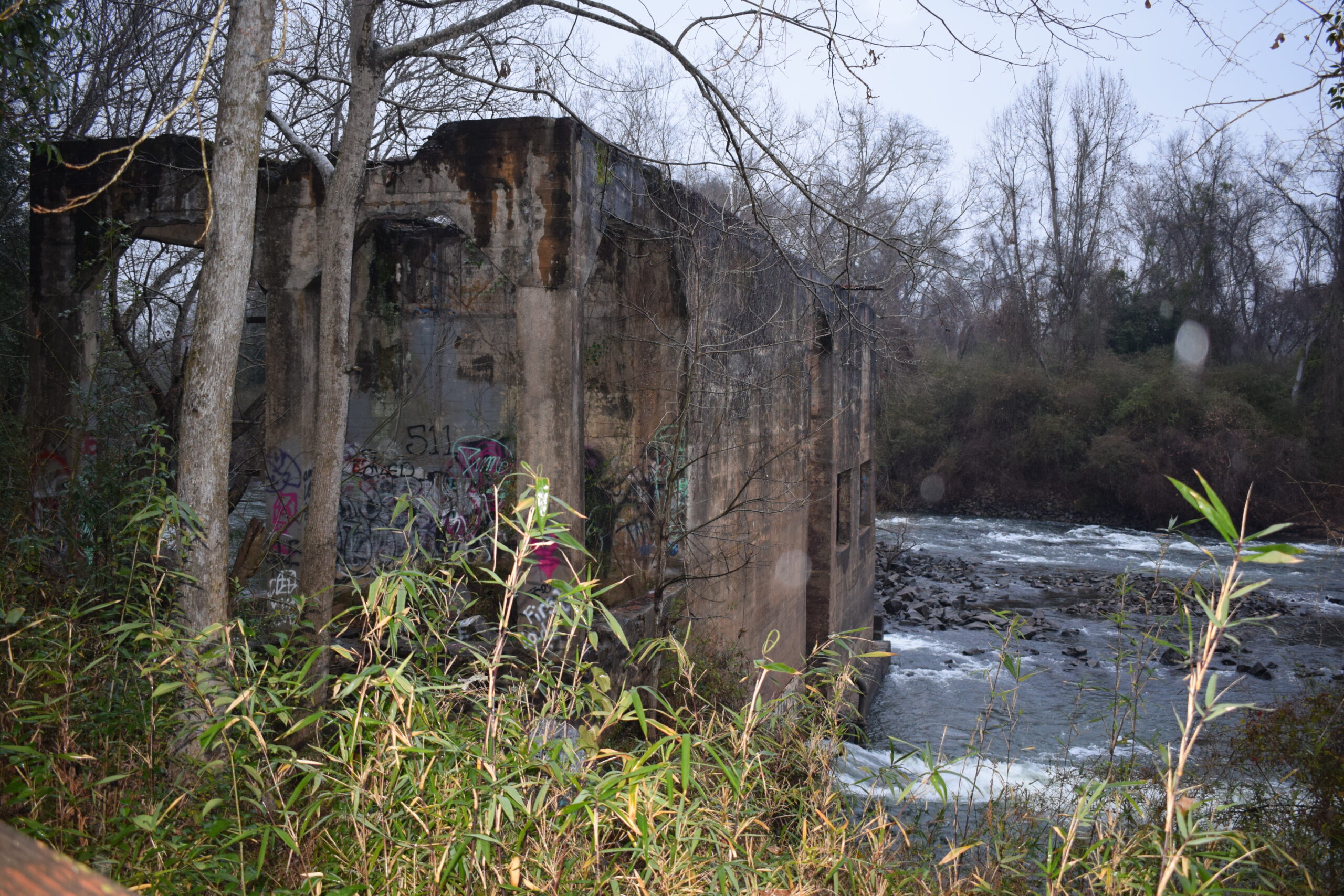 The weathered concrete ruins of the historic Oconee Mill stand along the rushing Oconee River near Milledgeville, Georgia, surrounded by winter trees and riverbank vegetation on a cold, rainy day.