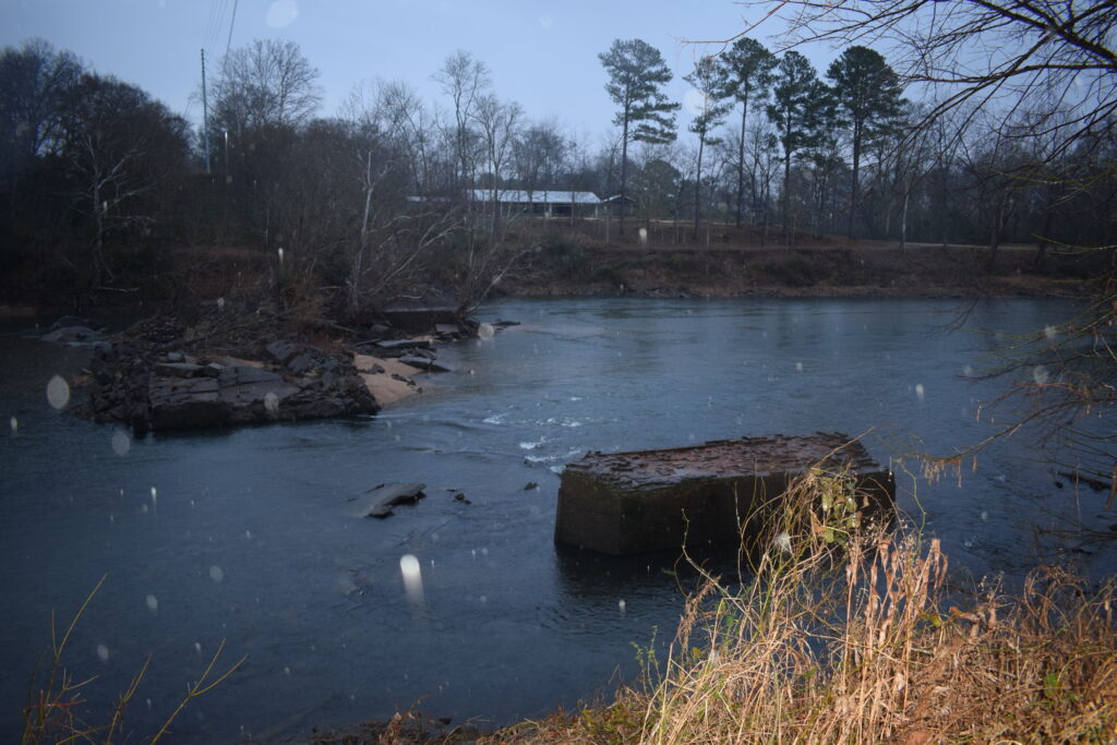 The Oconee River flows around scattered remnants of the historic Oconee Mill, with sections of the old foundation partially submerged in the water beneath a gray winter sky near Milledgeville, Georgia.