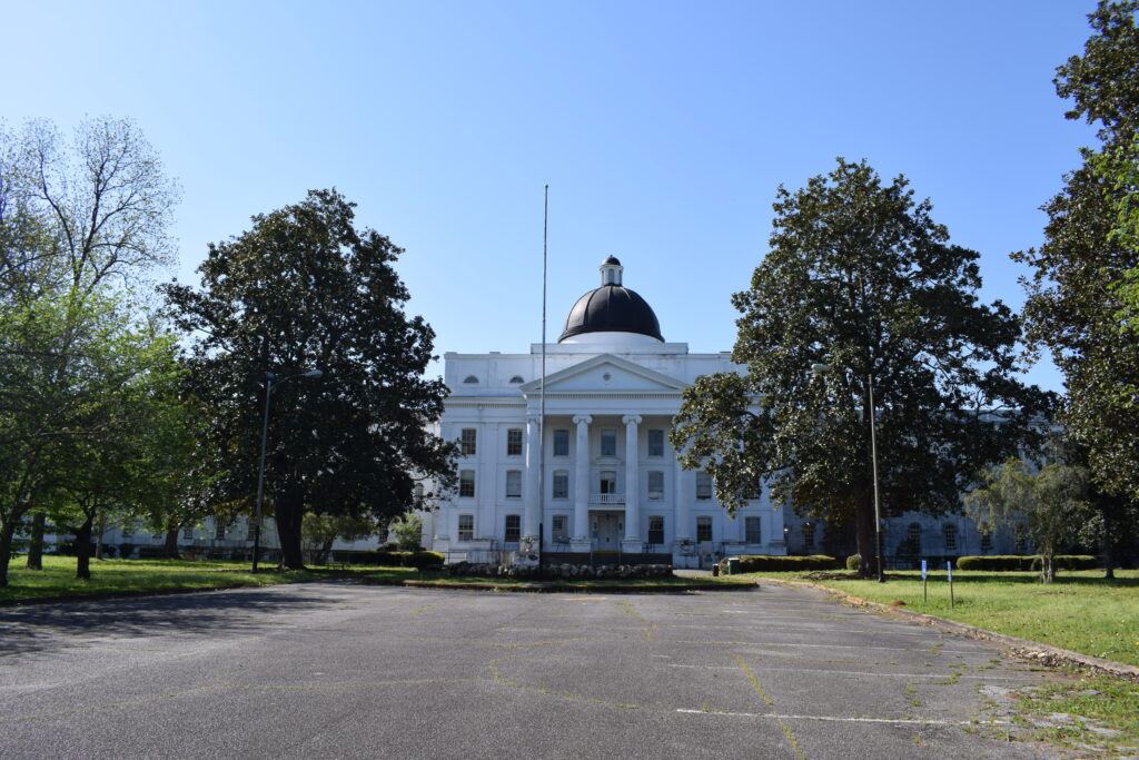 Front view of the historic Powell Building at Central State Hospital in Milledgeville, Georgia, framed by large magnolia trees beneath a clear blue sky.