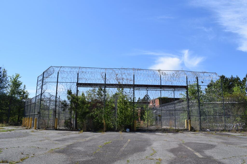 Wide view of the abandoned Scott State Prison perimeter with chain-link fencing, razor wire, and an empty parking lot in Milledgeville, Georgia.
