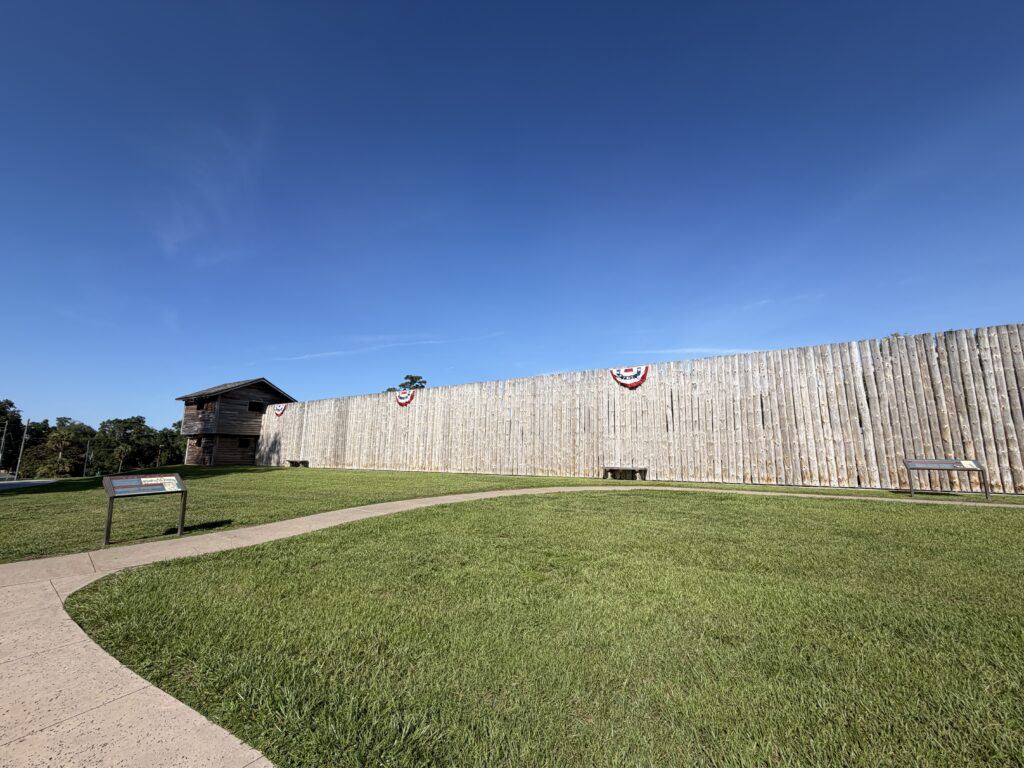 The reconstructed wooden palisade walls and blockhouse at Fort King National Historic Landmark in Ocala, Florida, stand beneath a clear blue sky with interpretive signs welcoming visitors to explore the historic fort.