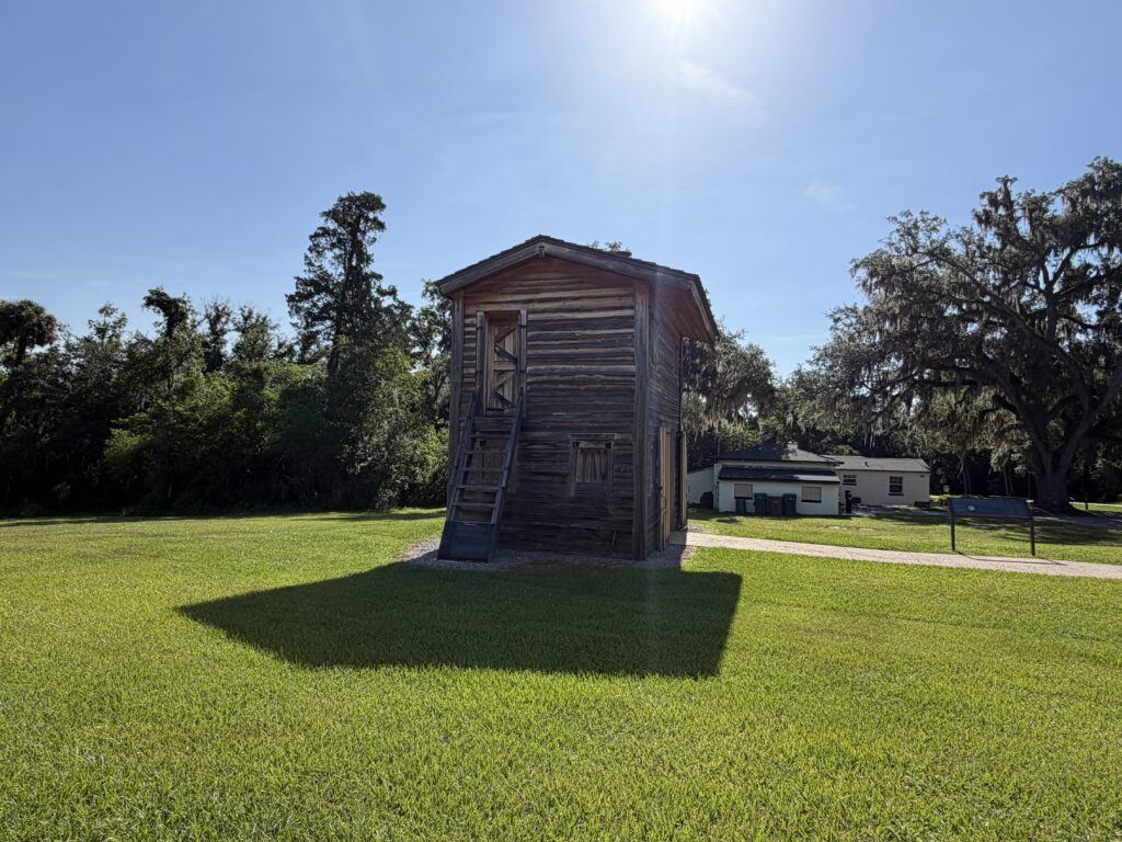 A reconstructed wooden blockhouse stands in the open parade grounds of Fort King National Historic Landmark in Ocala, Florida, surrounded by green grass, towering live oaks, and bright blue skies.