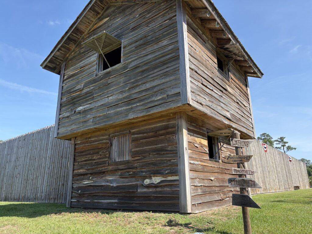 A reconstructed wooden blockhouse rises above the palisade walls at Fort King National Historic Landmark in Ocala, Florida, beneath a bright blue sky with directional signs pointing to historic features inside the fort.