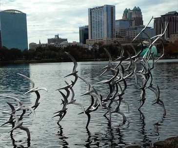 A metal sculpture of birds in flight stands along the shoreline of Lake Eola Park with the downtown Orlando skyline reflected across the water in the background.