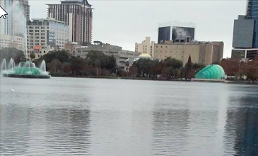 The iconic Lake Eola fountain stands before the downtown Orlando skyline, with the Walt Disney Amphitheater visible along the shoreline of Lake Eola Park.