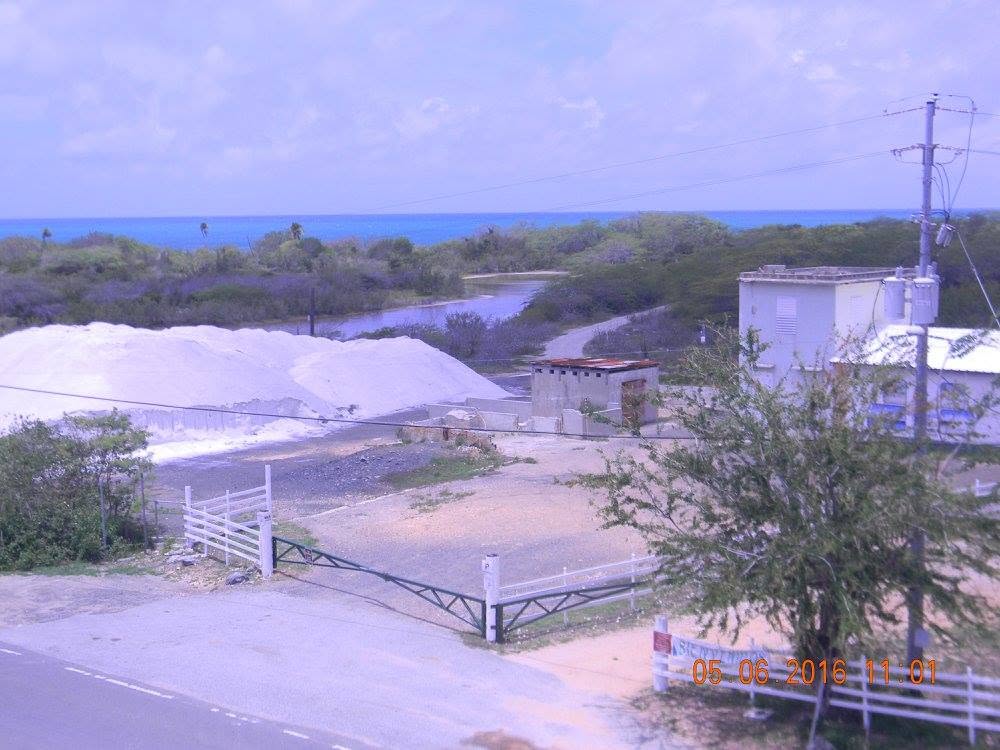 Mounds of harvested sea salt sit beside processing buildings at the Cabo Rojo Salt Flats in Puerto Rico, with the Caribbean Sea visible beyond the historic salt works.