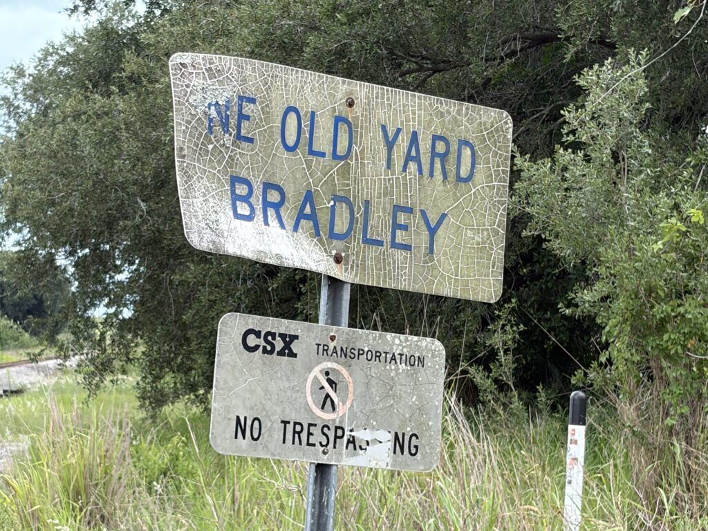 Weathered Ye Old Yard Bradley railroad sign beside CSX tracks in Bradley Junction, Florida, with a no trespassing sign below.
