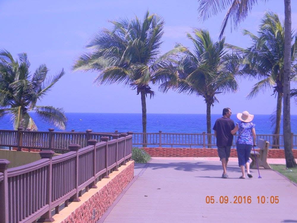 Visitors walking along the oceanfront path at Punta Higüero Lighthouse in Rincón, Puerto Rico, with palm trees and the Caribbean Sea beyond.