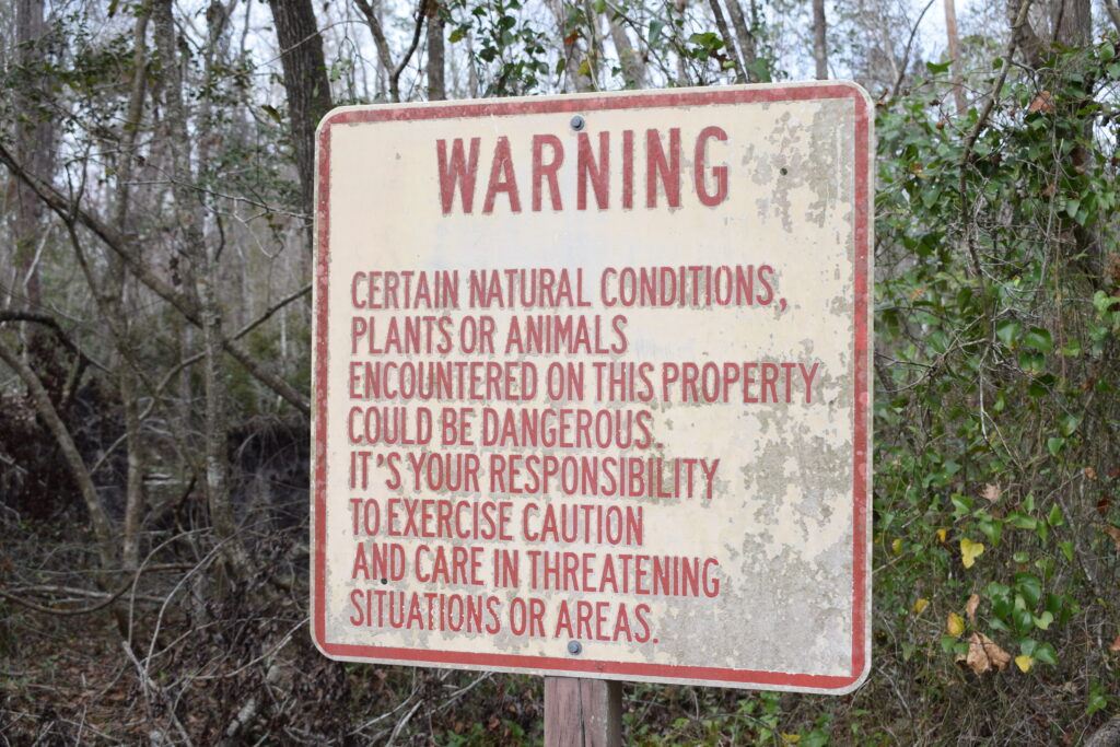 Weathered warning sign advising visitors about potentially dangerous natural conditions, plants, and animals at Falling Creek Falls in Florida.