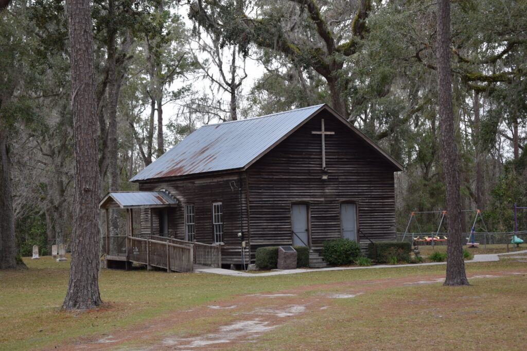 Historic wooden Falling Creek Church with cemetery graves visible beneath the trees near Lake City, Florida.