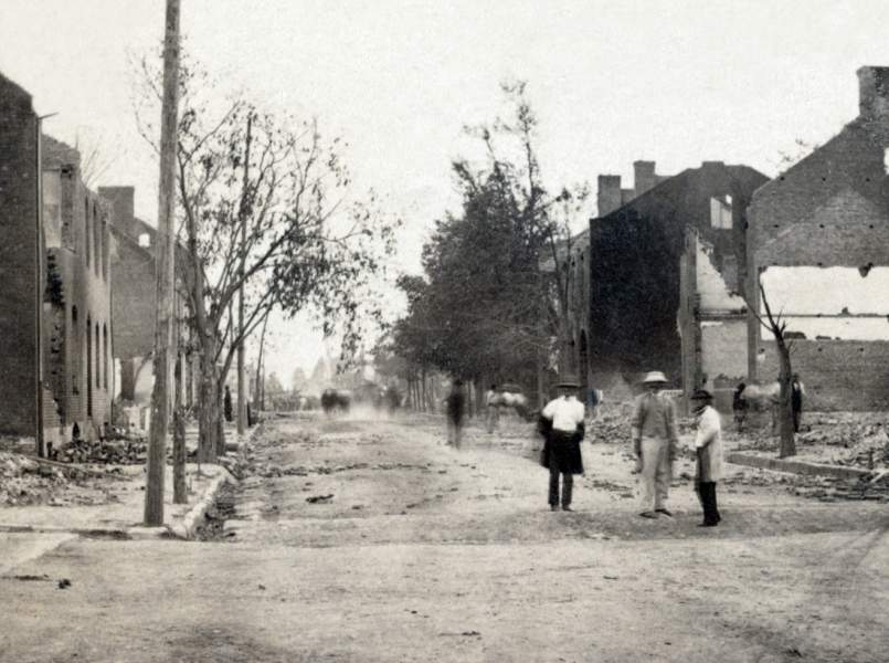 Historic black-and-white photograph showing a debris-covered street, burned-out buildings, and several men standing among the ruins of Chambersburg, Pennsylvania.
