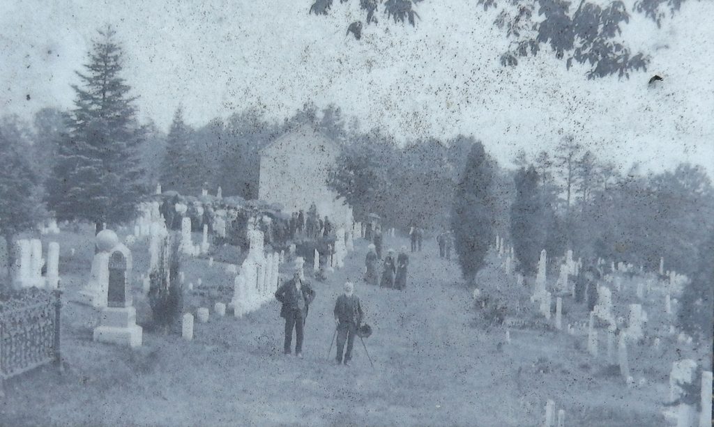 Historic black-and-white photograph showing two men standing among gravestones at Chestnut Ridge and Schellsburg Union Cemetery, with the Old Log Church visible in the background.