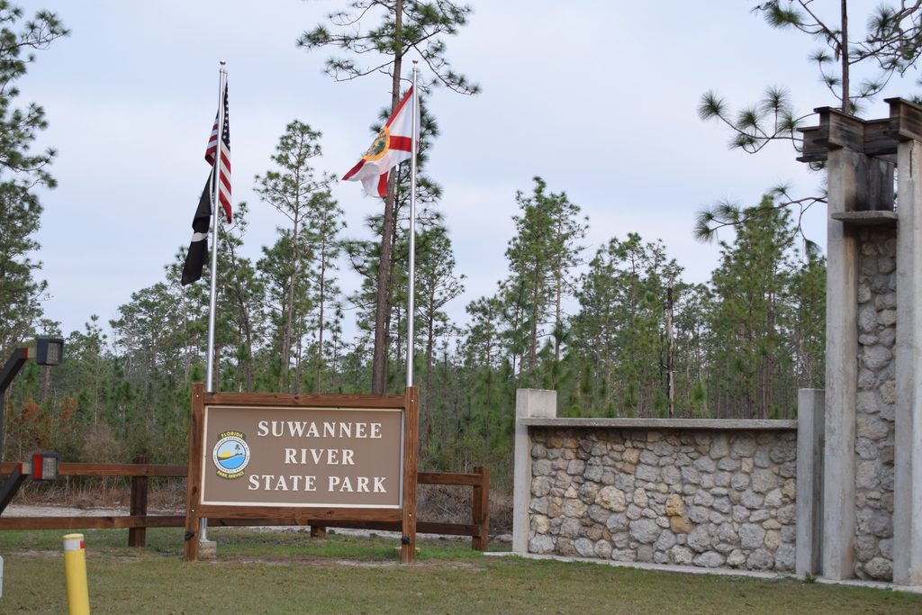 Entrance sign at Suwannee River State Park in Florida, with American and Florida flags, tall pine trees, and a stone entrance structure.