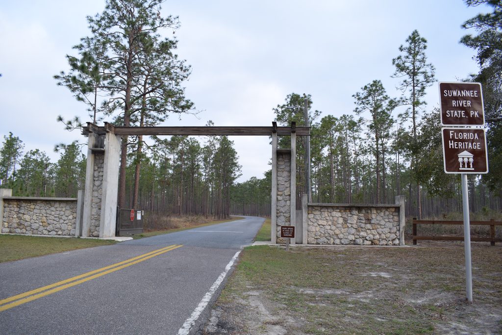 Rustic stone and timber entrance gateway to Suwannee River State Park, with a winding road, tall pine trees, and a Florida Heritage sign.