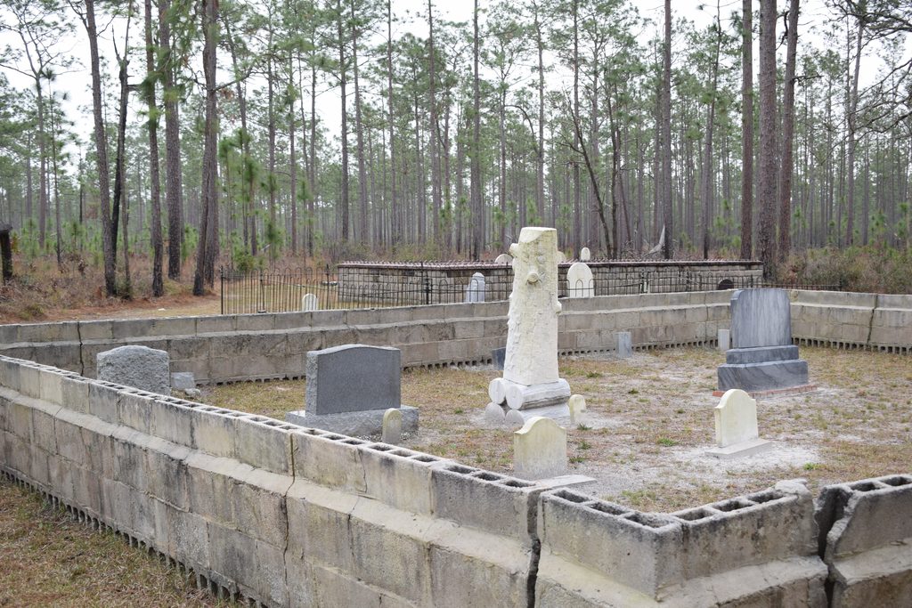 Historic gravestones, including a tall white tree-shaped monument, surrounded by low concrete block burial enclosures and pine forest at Columbus Cemetery in Suwannee River State Park.